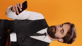 Vertical video Catering employee texting messages on phone while carrying restaurant tray with drinks and cutlery, posing in studio. Sophisticated waiter in suit and tie looking at smartphone display. - Powered by Shutterstock - Get 15% off with code: PIKWIZARD15