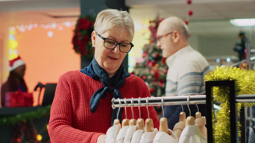 Elderly woman browsing through clothes in xmas adorn clothing store during winter holiday season. Senior client in shopping spree session at Christmas decorated fashion boutique in mall