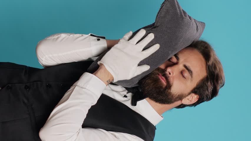 Vertical video Sleepy bellman posing with pillow in studio, advertising burnout and being overworked in classy hospital industry. Hotel porter taking quick nap after working overtime, tired doorman. Royalty-Free Stock Footage #1107481745
