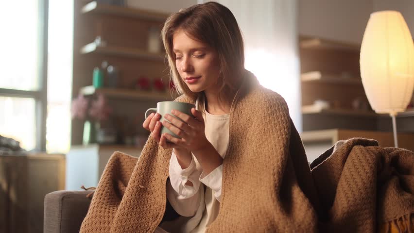 Portrait of charming blond young woman under the blanket take sip drinking hot tea or coffee sitting on couch at home Calm relaxed female enjoying weekend leisure time alone indoors Royalty-Free Stock Footage #1107483865