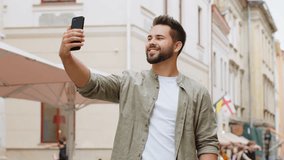 Bearded young man blogger taking selfie on smartphone, communicating video call online with subscribers or family friends, recording stories for social media vlog outdoors. Guy walking in city street