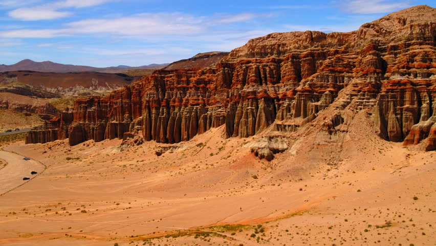 Aerial Panning Shot Of Red Rock Cliffs By Cars Near Mountain Ranges Against Sky - Red Rock Canyon State Park, California