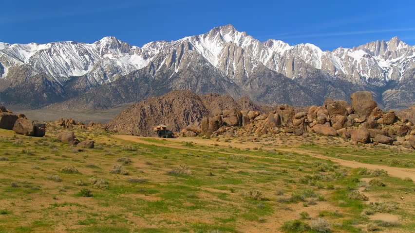 Aerial Forward Over Rocks By Tent And Car Near Snowcapped Mountains Against Blue Sky On Sunny Day - Red Rock Canyon State Park, California
