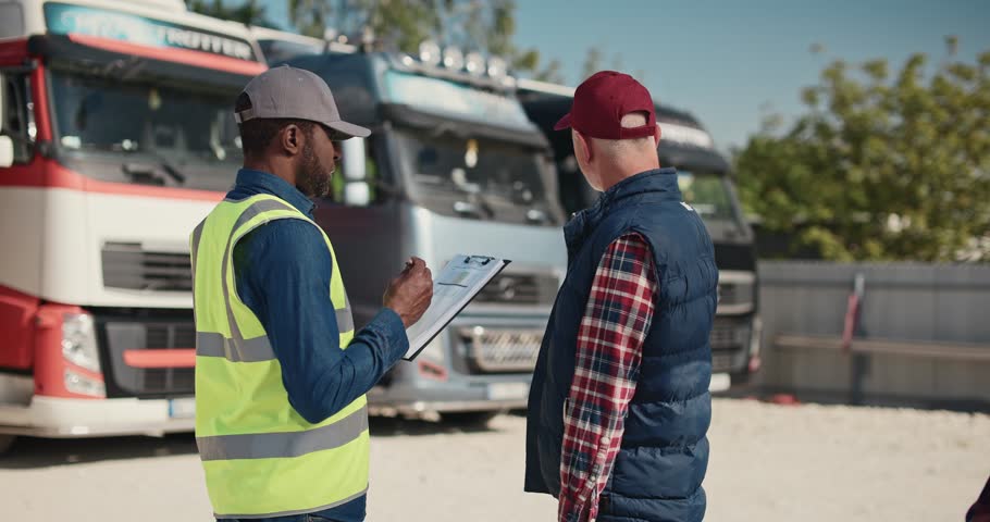 Side view of two multiethnic workers speaking with each other about load and unload freight at warehouse. Actively speaking while standing at lorries parking in sunny day. Shipping concept. Royalty-Free Stock Footage #1107486845