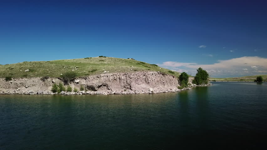 Aerial: Drone Flying Towards Green Hill With Trees Near Lake Against Blue Sky On Sunny Day - Billings, Montana