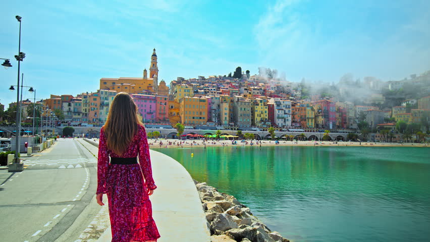 Panoramic detailed view of a beautiful girl walking towards colorful houses in Menton, France. Woman in a dress enjoys colorful houses and sand beach in the historical Old in Cote d'Azur, France. Royalty-Free Stock Footage #1107497335