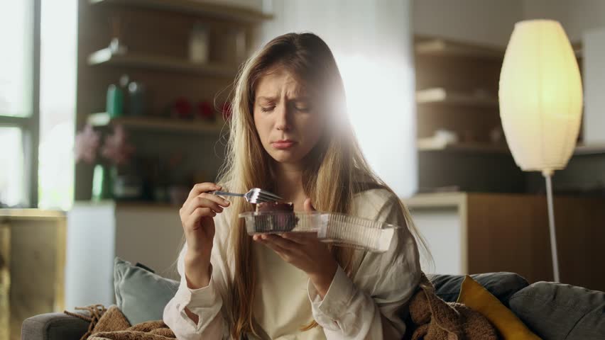Portrait of sad young woman dealing with stress by eating food sitting on sofa at home Upset female wrapped in blanket crying and eating cake indoors alone Mental heath problem Royalty-Free Stock Footage #1107503639