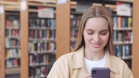 Happy pretty blonde girl student holding smartphone using mobile cell phone modern technology looking at cellphone standing in modern university campus library checking tech device.