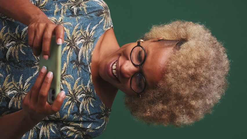 Vertical video, Happy girl in glasses dressed in blue t-shirt, holds mobile phone in her hands, smiles and plays games on mobile phone isolated on green-blue background