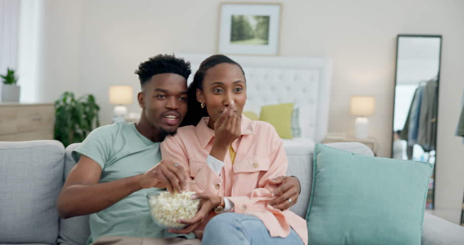 Black couple on sofa with popcorn, watching tv comedy and date night together in living room. Relax, African man and happy woman eating snacks on couch with laughing, comic show and funny movies. Royalty-Free Stock Footage #1107519359
