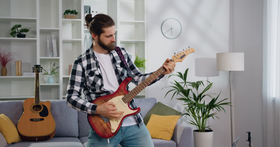 Handsome bearded man musician plays the guitar, takes pleasure of music and his hobby at ligt living room. Modern entertainment, music and happy young people concept. Slow motion.