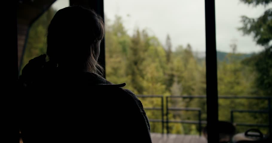 A happy blonde girl with a cup of coffee goes to the balcony of a country house and looks at the lush green coniferous forest in the mountains Royalty-Free Stock Footage #1107521239