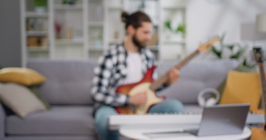 Portrait of young bearded man learning to play music instrument and looking at camera. Handsome musician sitting on sofa playing electric guitar in light living room and posing to camera