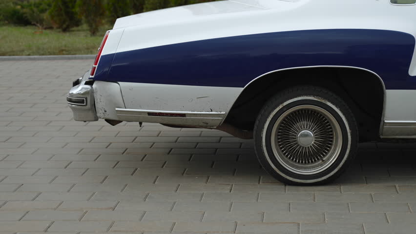 Back of vintage car with engine running and gases coming out of exhaust pipe. Beautiful spoked rims on cloudy day and non-environmental exhaust from engine of old blue and white car.