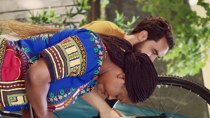 Vertical video Vibrant african american lady browsing on internet for bicycle gear maintenance. Black woman checking for repair instructions on laptop while caucasian man dismantles broken bike wheel. | Shutterstock HD Video #1107526739