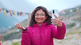 Young beautiful Indian girl tourist in warm jacket making video call with smartphone on mountain background. Happy woman talking video with friends. Vacation and holidays concept