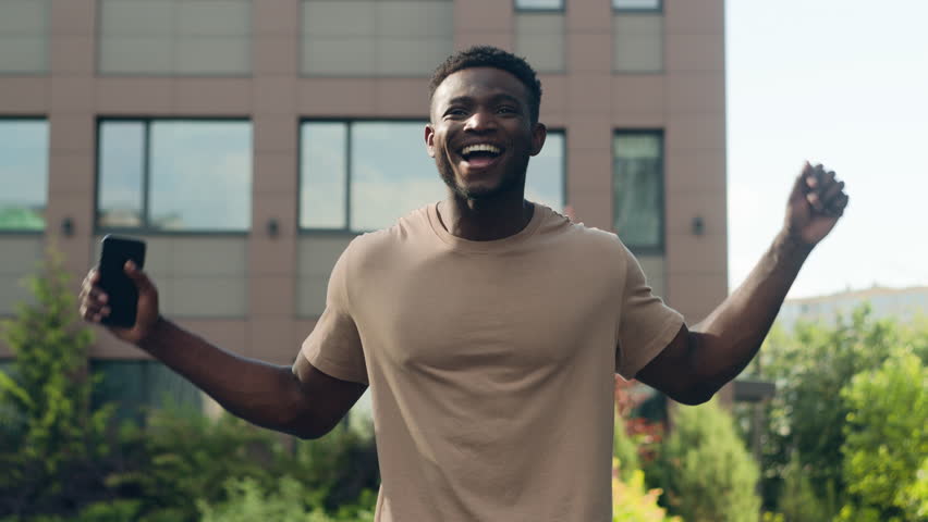 African American happy success winner man ethnic guy in city male outdoors win victory achieve with mobile phone smartphone achievement jump scream yelling good news rejoice jumping champion gesture Royalty-Free Stock Footage #1107529647