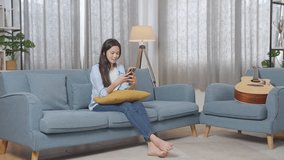 Full Body Of Asian Teen Girl Enjoys Playing Smartphone While Sitting On Sofa In The Living Room At Home
