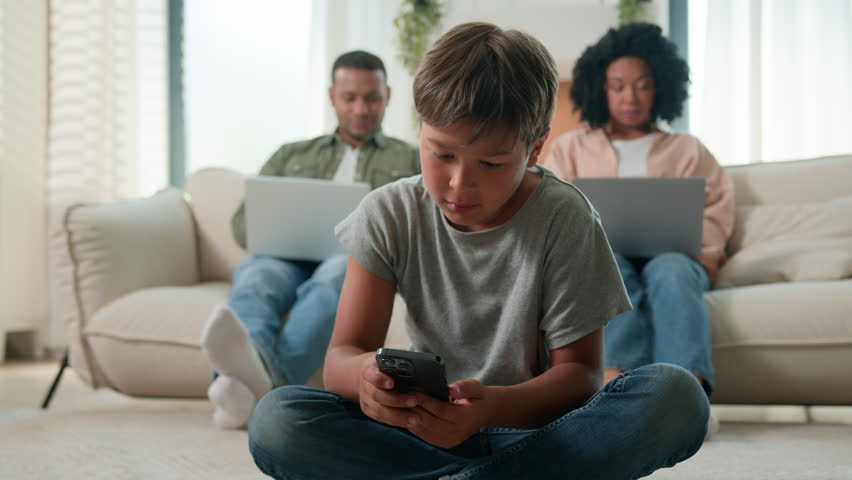 Modern gadgets addicted family using diverse devices at home African American multiracial parents on couch working online with laptops together little boy kid son child play video game on mobile phone