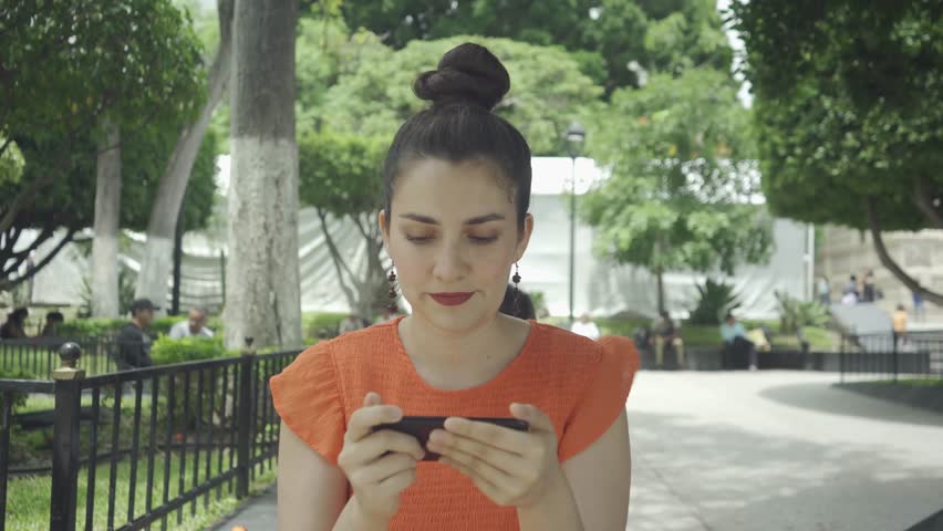 Young Latin woman is happy watching movies or videos on his phone at the park making face expressions. Daylight shooting in downtown of a city in Mexico, Latin America. SHOTLISTstream