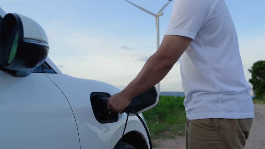 Asian man insert smart EV charger to electric car powered by wind turbine. Renewable energy utilization for sustainable clean EV technology with futuristic digital battery status hologram. Peruse