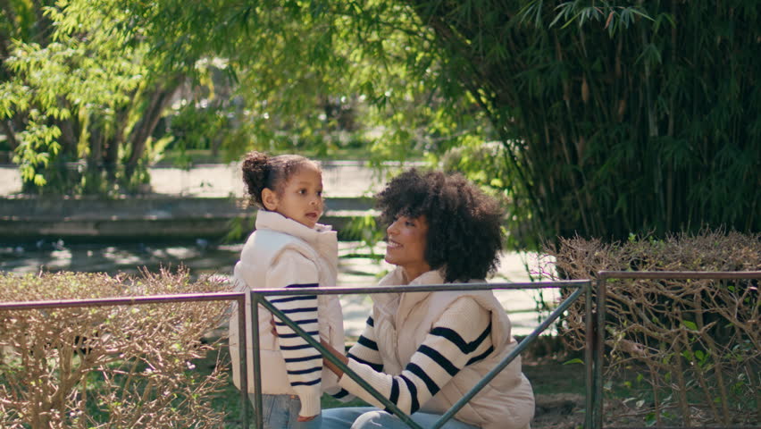Woman carrying daughter on walk transplanting over fence. Cheerful african american mother taking little girl on hands strolling at sunny park together. Happy mom have fun on nature with cute child.