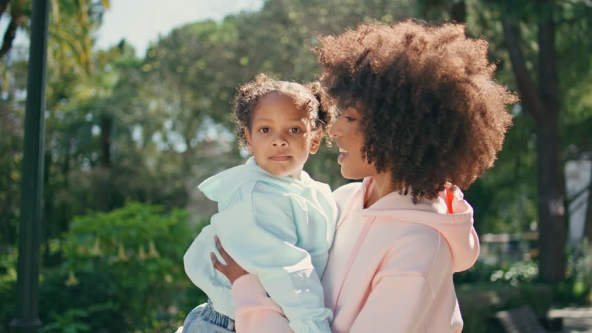Mother standing with cute daughter on hands enjoying motherhood close up. Happy smiling african american woman holding little girl relaxing in sunny park. Curly cheerful mom feeling love to cute child