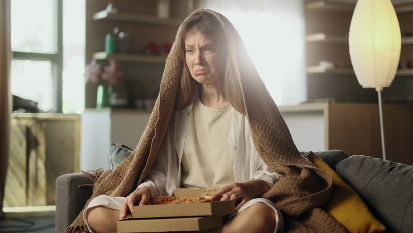 Portrait of sad young woman dealing with stress by eating food sitting on sofa at home Upset female wrapped in blanket crying and eating pizza fast food indoors alone Mental heath problem Royalty-Free Stock Footage #1107560253