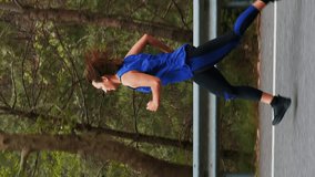 Female running on empty asphalt road in forest. Vertical video. Woman doing sport exercises outdoor for healthy lifestyle. Sport training. Concentrated young female athlete practicing in forest