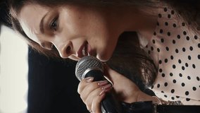 Female singer singing song into microphone. Vertical video. Portrait of young charming woman singing lyrics at sound recording studio. Professional vocalist practicing at rehearsal studio