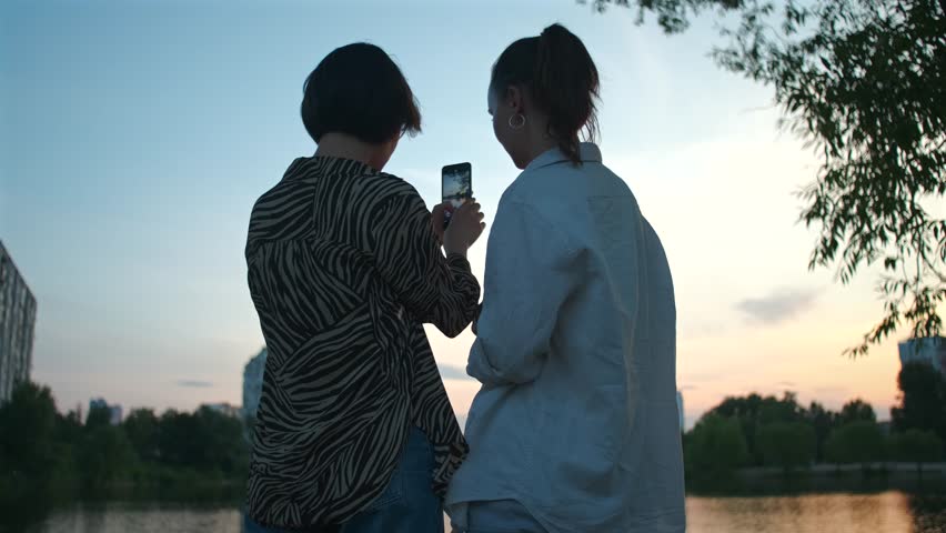 Two women take photos on camera. Beautiful girls taking video of sunset and lake on mobile phone. Couple of female friends in warm summer evening, nature landscape Royalty-Free Stock Footage #1107575393