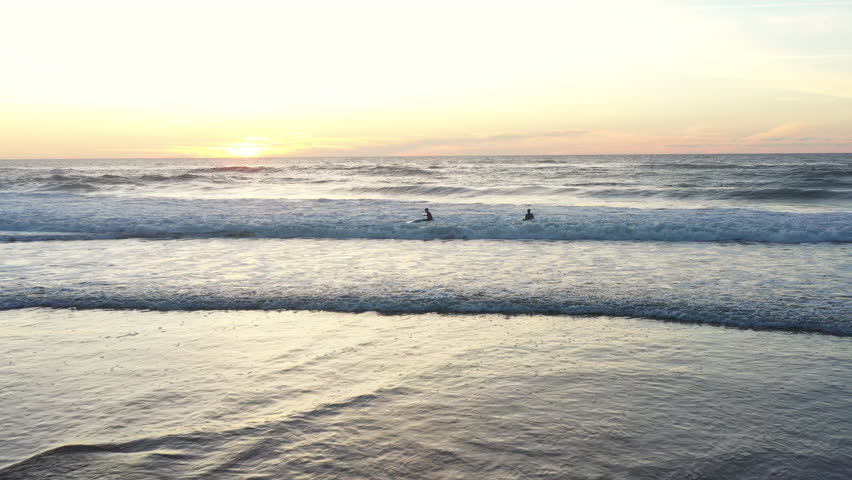 Two surfers are training to ride waves in Atlantic Ocean at sunset wearing wetsuits. Aerial view