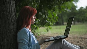 young beautiful woman with a laptop in her hands talking via video link using a webcam sitting on lawn near a tree in park