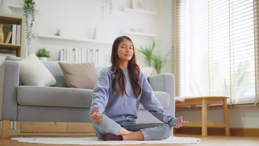 Happy young Asian woman practicing yoga and meditation at home sitting on floor in living room in lotus position and relaxing with closed eyes. Mindful meditation and wellbeing concept Royalty-Free Stock Footage #1107594611