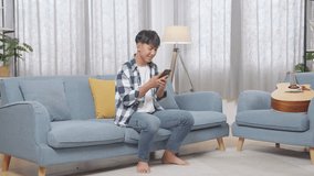 Full Body Of Asian Teen Boy Enjoys Playing Smartphone While Sitting On Sofa In The Living Room At Home
