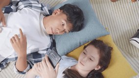 Close Up Top View Of Asian Teen Couple Having Video Call On Smartphone While Lying On Carpet On The Floor At Home. Waving Hands, Laughing, And Speaking
