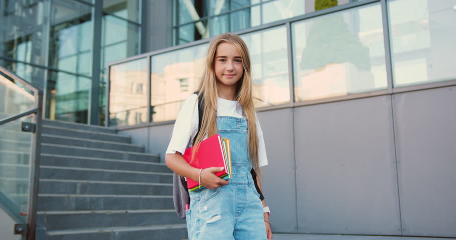 Portrait of lovely teen school girl with backpack smiling posing on camera near campus. Female elementary school student holds textbooks standing on stairs outside school building. Royalty-Free Stock Footage #1107605803