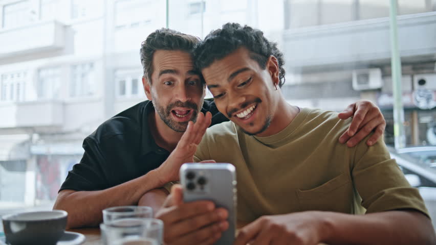 Couple gays greeting at video call by smartphone sitting comfortable cafeteria close up. Two multiethnic men waving hands to phone camera. Smiling homosexual partners talking at online meeting in cafe Royalty-Free Stock Footage #1107610443
