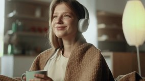 Portrait of pretty young woman under the blanket listening to music on sofa with soft light behind her indoors Smiling female with headphones hold cup of hot drink enjoying leisure time at home alone - Powered by Shutterstock - Get 15% off with code: PIKWIZARD15