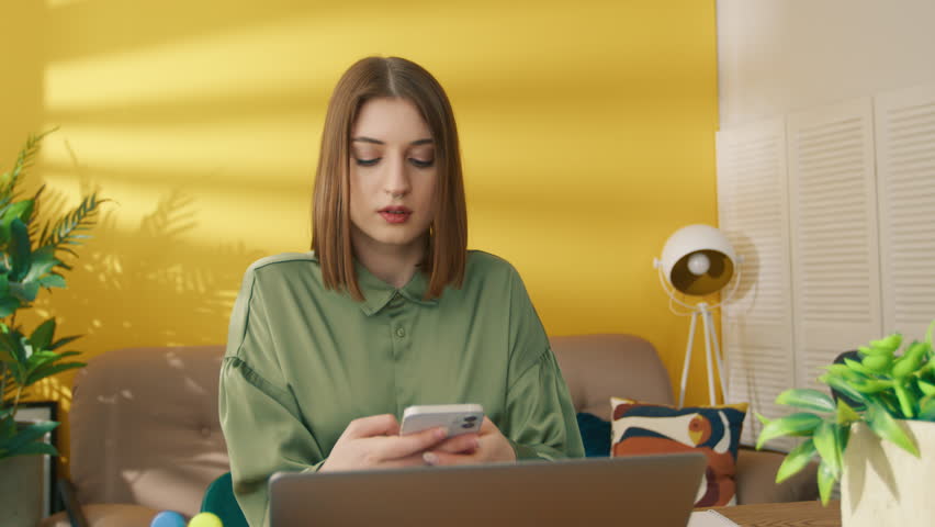 Serious woman learning online on laptop at home. Focused girl reading message on smartphone. Concentrated lady typing on mobile phone. Smart student taking break between online learning, 4k footage