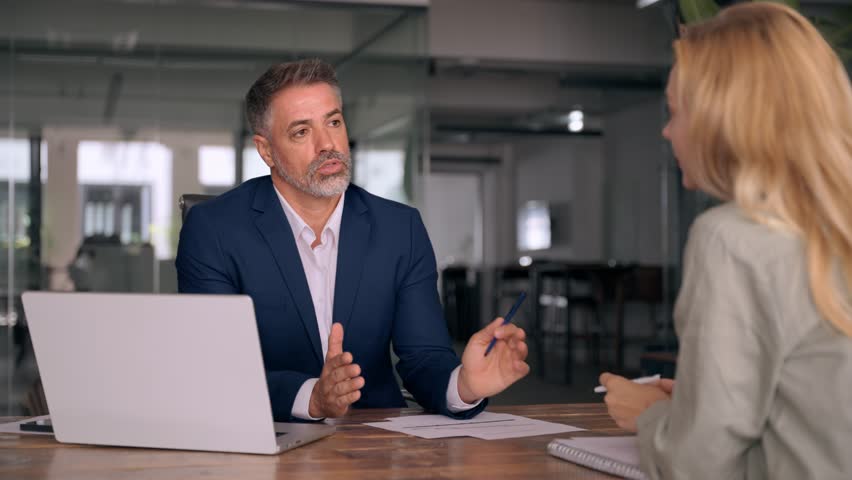 Lawyer or real estate agent make consultation for client. Mature Latin business man and European business woman discussing project on laptop sitting at table in office. Business people work together. Royalty-Free Stock Footage #1107618543