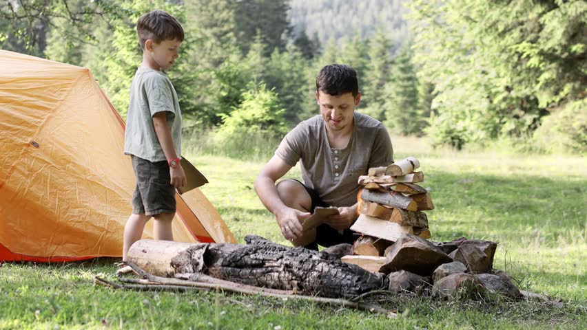 Dad and son sit together by a bundle of firewood and light a fire near the tent. The spirit of adventure and adventure. Hiking and camping. Travel concept 4K.
