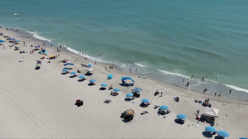 aerial footage of a gorgeous summer landscape at the beach with people relaxing in the sand under colorful umbrellas with ocean water, waves in Carolina Beach North Carolina USA