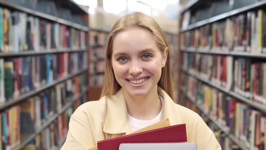 Happy pretty blonde european teen girl, smiling female teenage high school student holding notebooks looking at camera standing in modern university or college campus library, portrait. Royalty-Free Stock Footage #1107621901