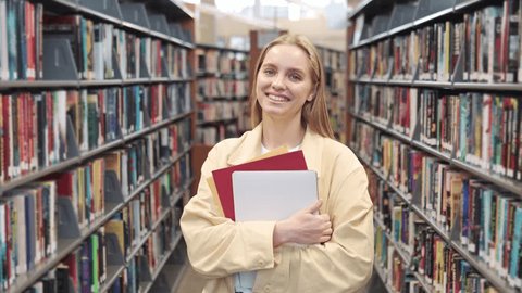 Happy pretty blonde european teen girl, smiling female teenage high school student holding notebooks looking at camera standing in modern university or college campus library, portrait. 库存视频