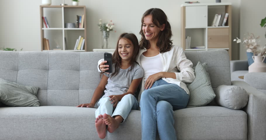 Positive young mother and pretty preschool girl using online media application on smartphone, taking funny selfie on mobile phone, self video for social network, watching content on Internet Royalty-Free Stock Footage #1107623905