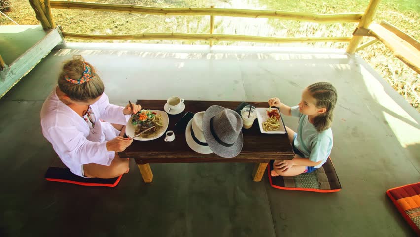 The happy family eat and drink tasty beverages spending time in local floating cafe on water. Mother and daughter having breakfast outdoors. Woman and a child in a cafe. Tropical view