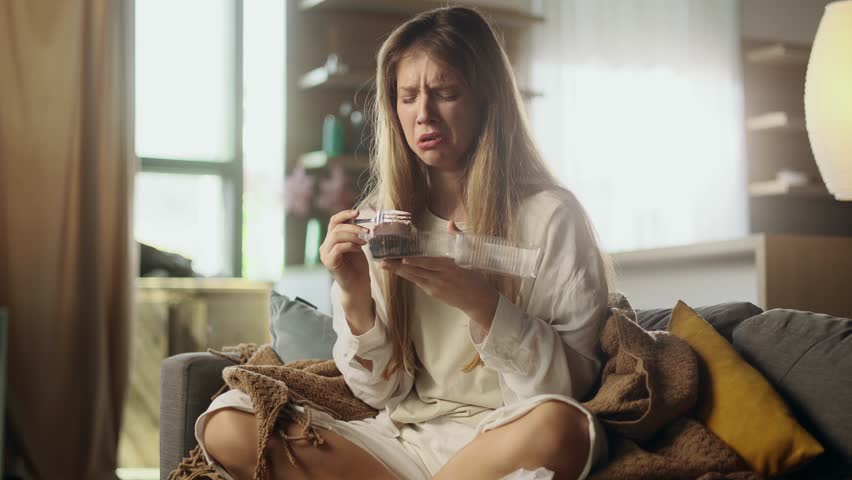 Portrait of sad young woman dealing with stress by eating food sitting on sofa at home Upset female wrapped in blanket crying and eating cake indoors alone Mental heath problem Royalty-Free Stock Footage #1107635539
