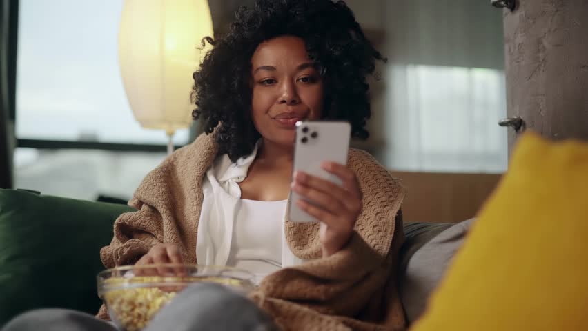 Charming young curly woman with bowl of popcorn hold smartphone browsing scrolling app watching social media feed indoors Happy female texting on her phone enjoying leisure time at cozy home Royalty-Free Stock Footage #1107635579