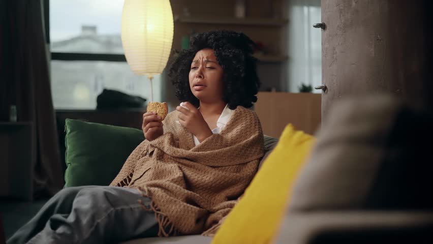 Portrait of sad young woman dealing with stress by eating food sitting on sofa at home Upset female wrapped in blanket crying and eating cookies indoors alone Mental heath problem Royalty-Free Stock Footage #1107635583